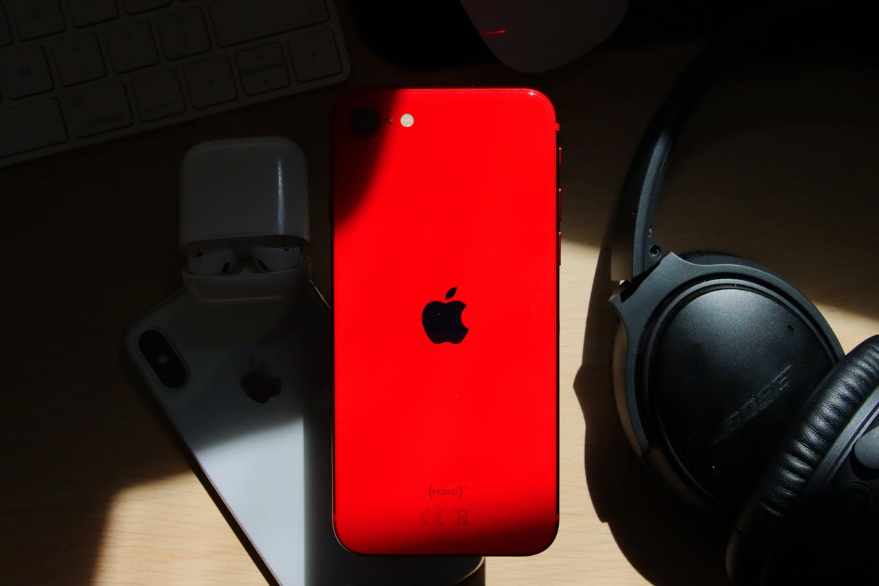 A flat lay of a red iPhone surrounded by AirPods and Bose headphones on a wooden surface.